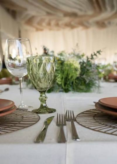 Mesa de boda bajo carpa con copas de cristal verde, vajilla terracota y camino de follaje