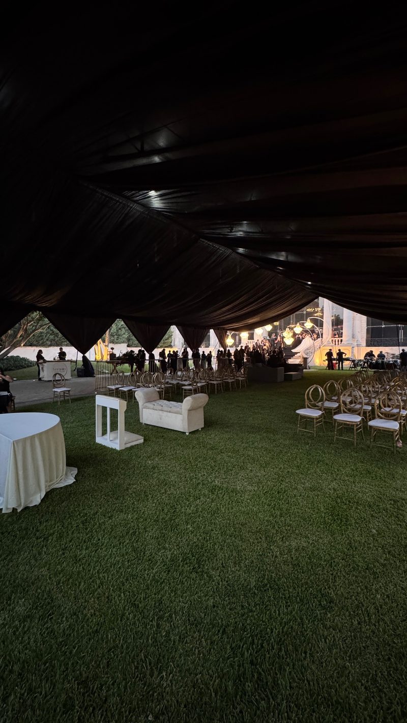 Ceremonia montada bajo carpa en jardín con sillas doradas y salón iluminado al fondo