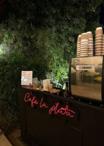 Barra de café con máquina profesional, vasos y letrero de neón, montada sobre muro verde en un evento nocturno