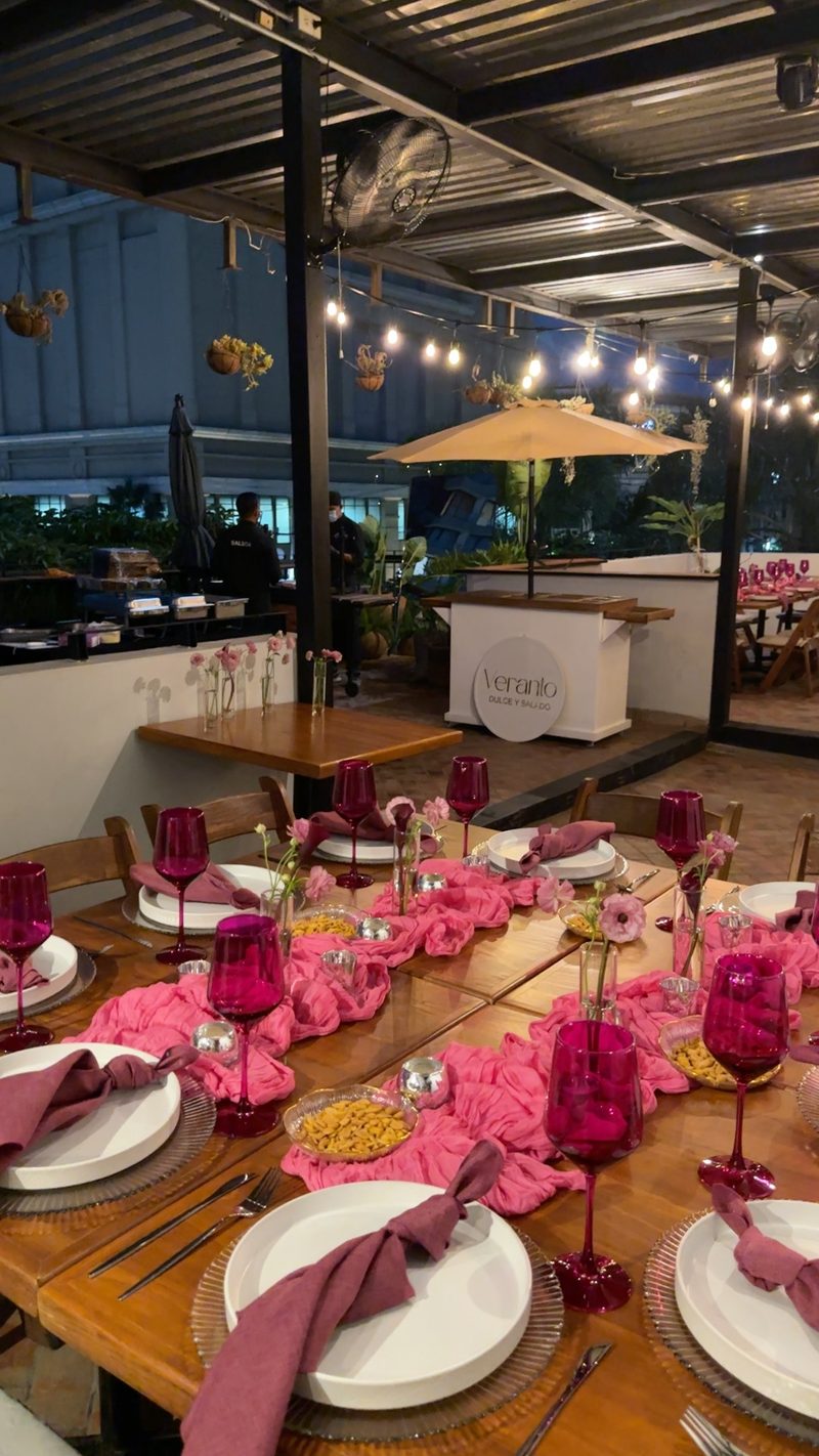 Mesa larga en terraza con mantelería rosa, copas magenta y luces colgantes