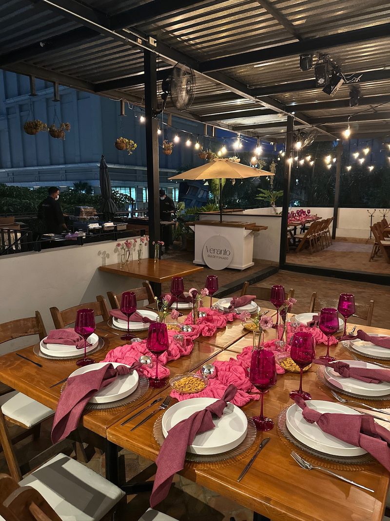 Vista amplia de terraza con mesas en tonos rosa, copas magenta y luces colgantes